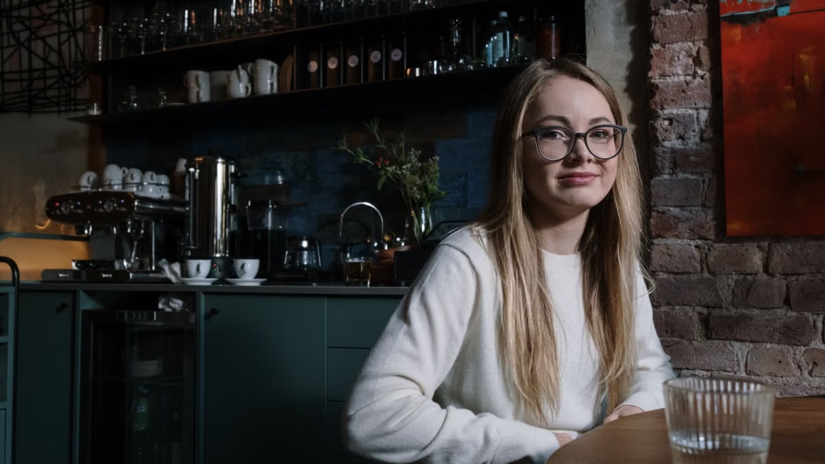 A smiling young woman with eyeglasses sitting in a stylish coffee shop interior.